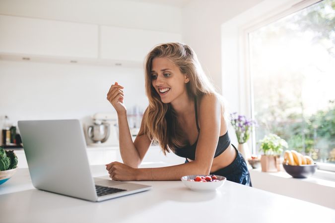 Woman in shorts and sports bra leaning on the kitchen counter with laptop