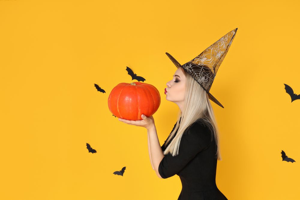 A woman in a wizard's hat kisses a pumpkin, on a yellow background
