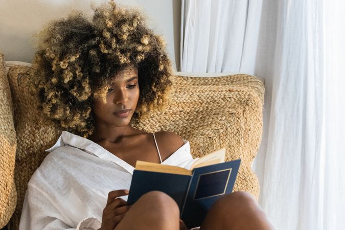 Young Black woman sitting on bed and reading book