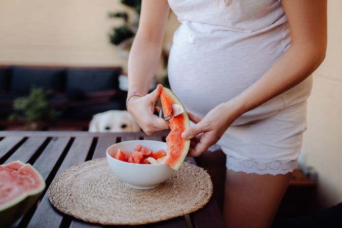 Faceless pregnant woman cutting watermelon to bowl
