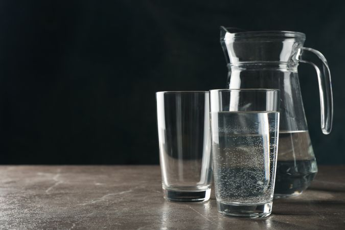 Two glasses of water in dark room on marble table with pitcher, copy space