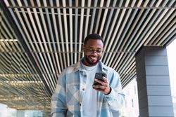 Cheerful Black man browsing smartphone near modern building