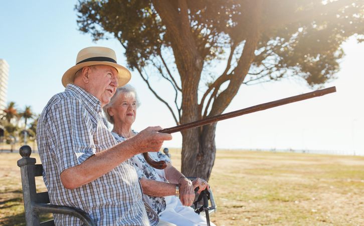 Side view of older couple relaxing on a park bench with man pointing out