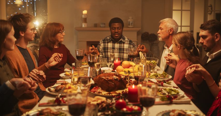 Portrait of a Young Black Man Sitting at a Festive Table with Family and Friends, Praying Before Eating a Turkey Dinner.
