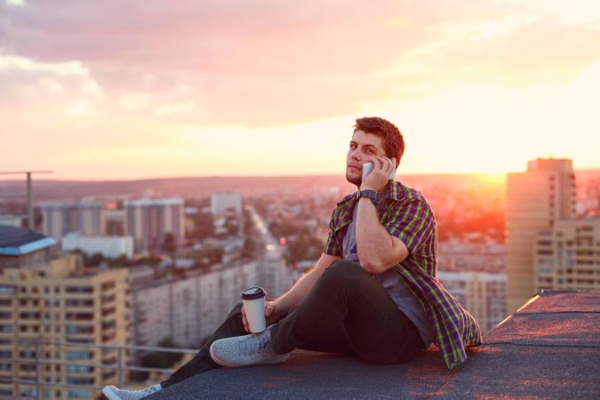 Male taking phone call while sitting on roof with to go coffee
