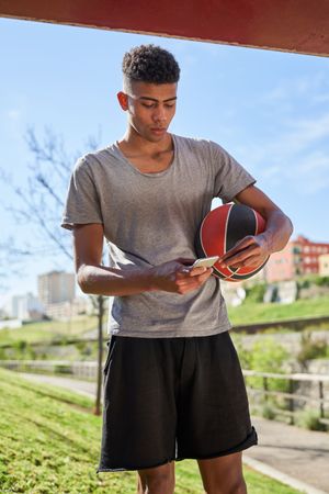 Latino fit man with ball using smartphone on sunny day