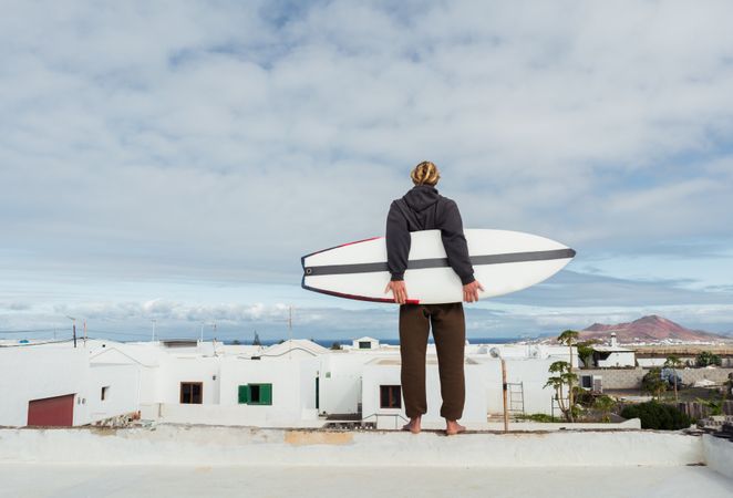 Man standing with surf board on roof of building and looking at mountain