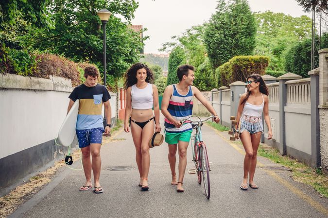 Happy young people walking along road in summer day with surfboard and bicycle