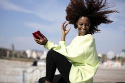 Carefree smiling female with curly hair in green shirt sitting listening to music on dock