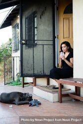 Woman sitting on front steps of house holding glass and dog laying next to her 5oD884