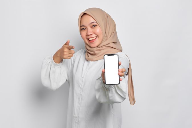 Asian Muslim woman in a bright studio shoot holding cell phone and pointing forward