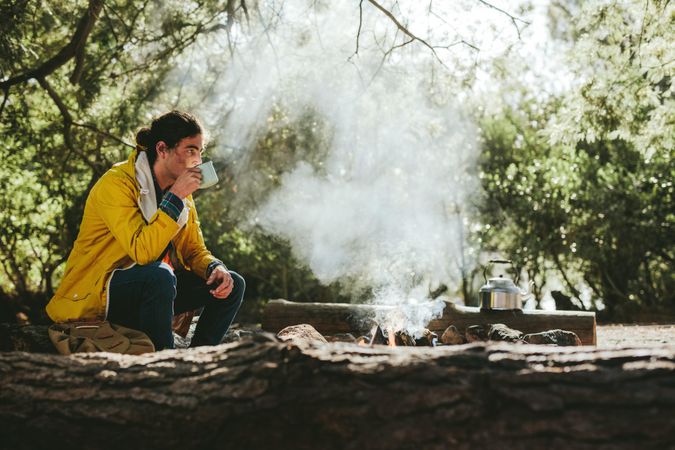 Young hiker enjoying a cup of tea sitting beside a campfire in a forest