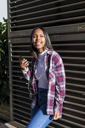 Female leaning on wall outside on sunny day listening to music on smartphone