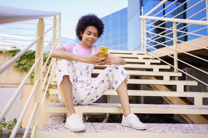 Woman in floral overalls sitting on stairs checking phone