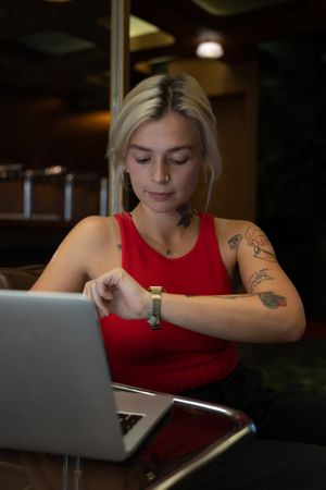 Young woman in red tank top working on laptop computer and looking at her watch in a coffee shop