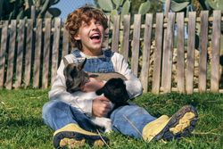 Joyful boy hugging cute baby goats and laughing sitting on lawn