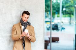 Man leaning on cement wall outside and wearing a camel coat while looking down at phone smiling