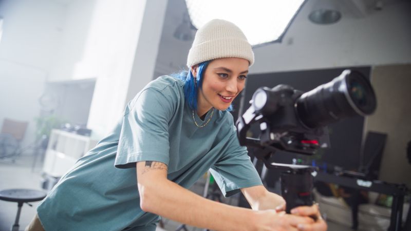 Young Female Cinematographer With Blue Hair Adjusts Professional Camera In A Bright Studio Setting, Focusing Intently On Capturing Creative Content For Film Or Advertising Production.