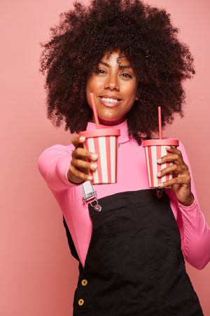 Happy Black woman with disposable cup of drink standing against pink background