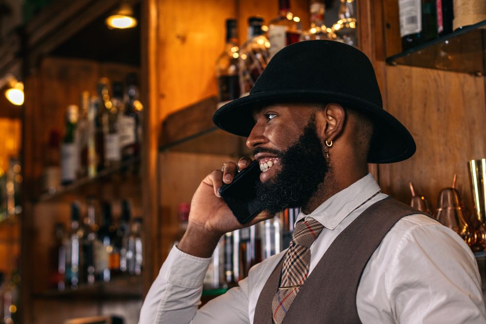 Stylish Black bartender in hat using smartphone