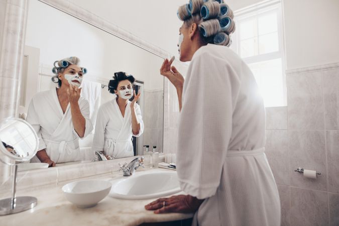 Two women with curling rollers to hair applying cream on face at home in the bathroom