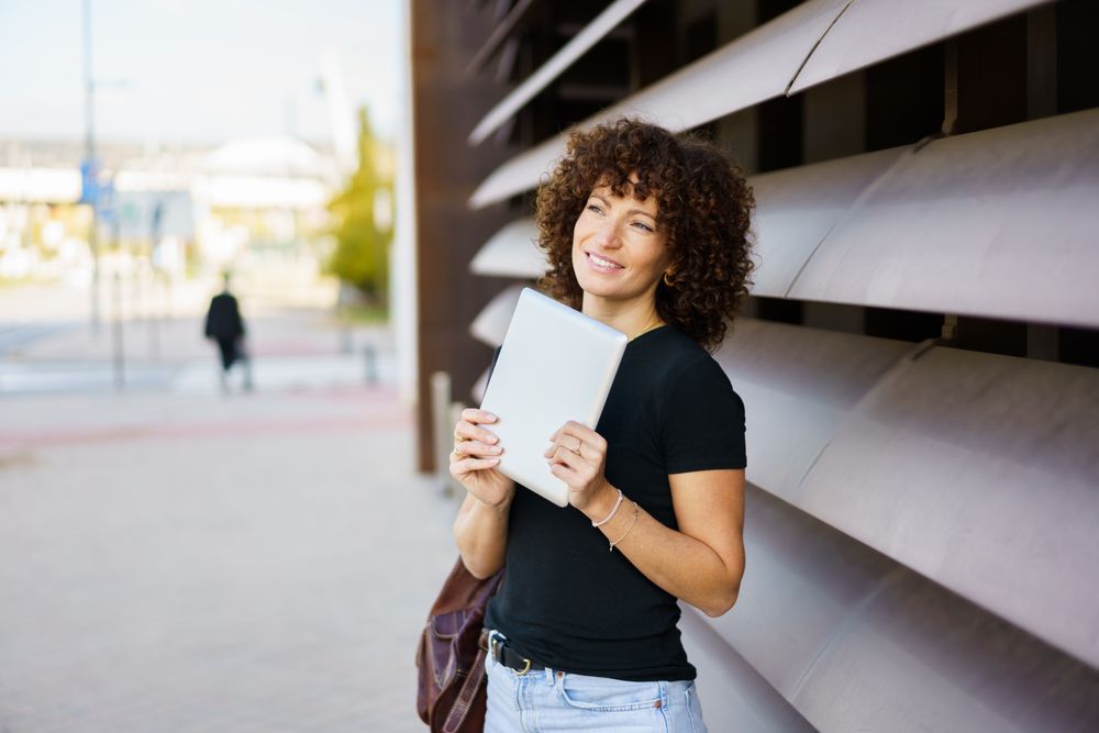 A Confident Young Woman Holding a Tablet While Outdoors in a Modern Urban Setting