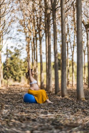 Adult pregnant lady sitting on big pilates ball in park during sunny day