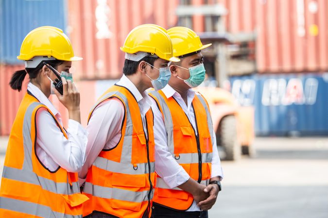 Three people in PPE gear on dock with shipping containers