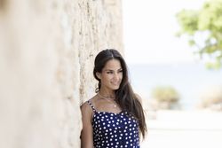 Latina woman in blue dotted dress leaning on wall outside and looking away
