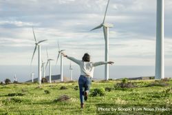 Back view of excited woman in light sweater and denim pants standing beside wind turbines 5awRWb