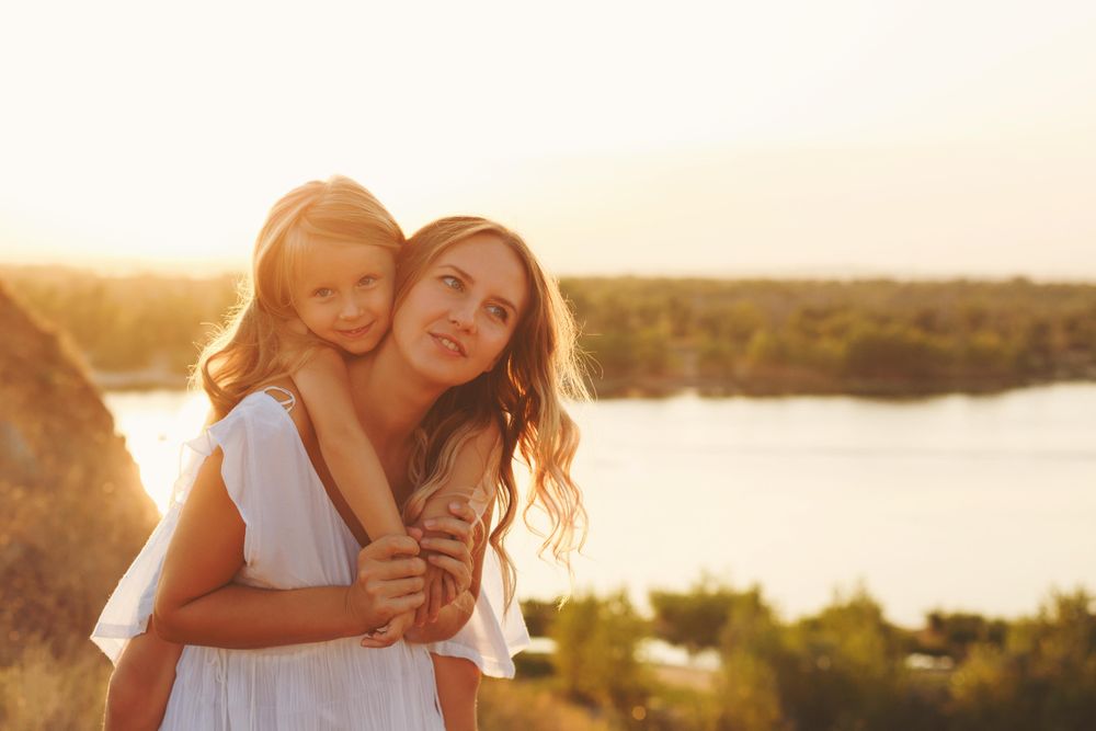 Young happy girl on woman’s back in front of river at dusk