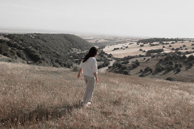 Back view of woman in light shirt and gray pants standing against bushes on hills