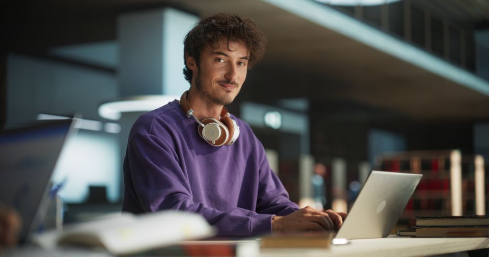 Portrait of a European Student Studying in a Traditional Library. Young Male Looking at Camera and Smiling. Scholar Working on Laptop Computer in College, Researching Academic Topics Online