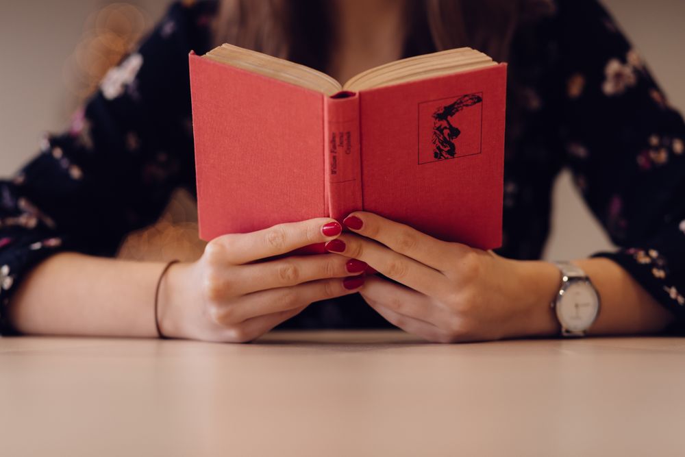 Cropped image of woman sitting at a table reading a book with red cover