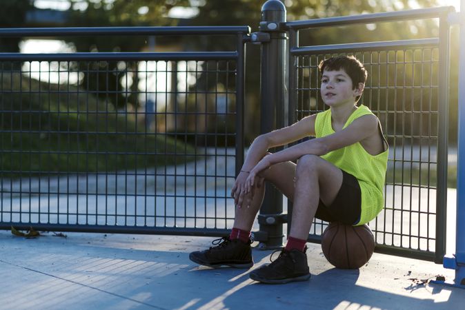 Young male outdoors sitting on basketball at a public court