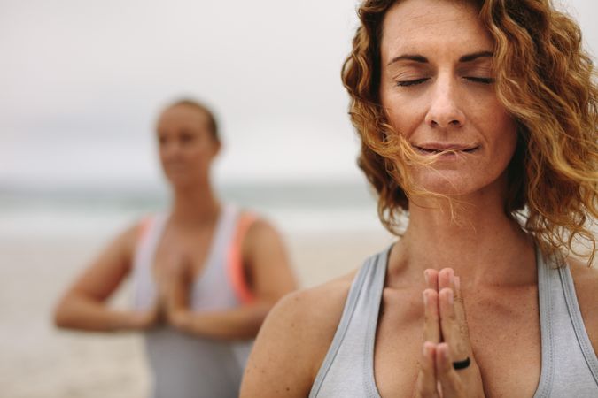 Two fit women sitting with prayer hands outdoors near ocean