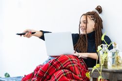 Woman with dreadlocks working with a laptop at home and with a remote control in hand
