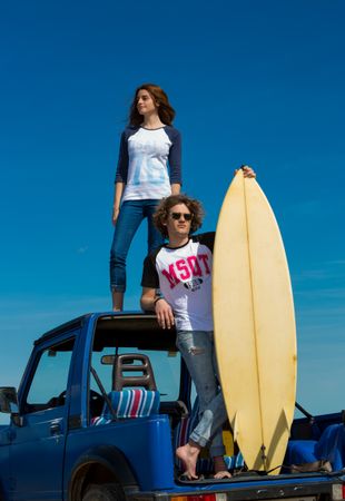 Guy with surfboard and woman on roof of car