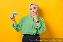 Muslim woman in headscarf and green blouse holding credit card and making surprised face 0JBmw0