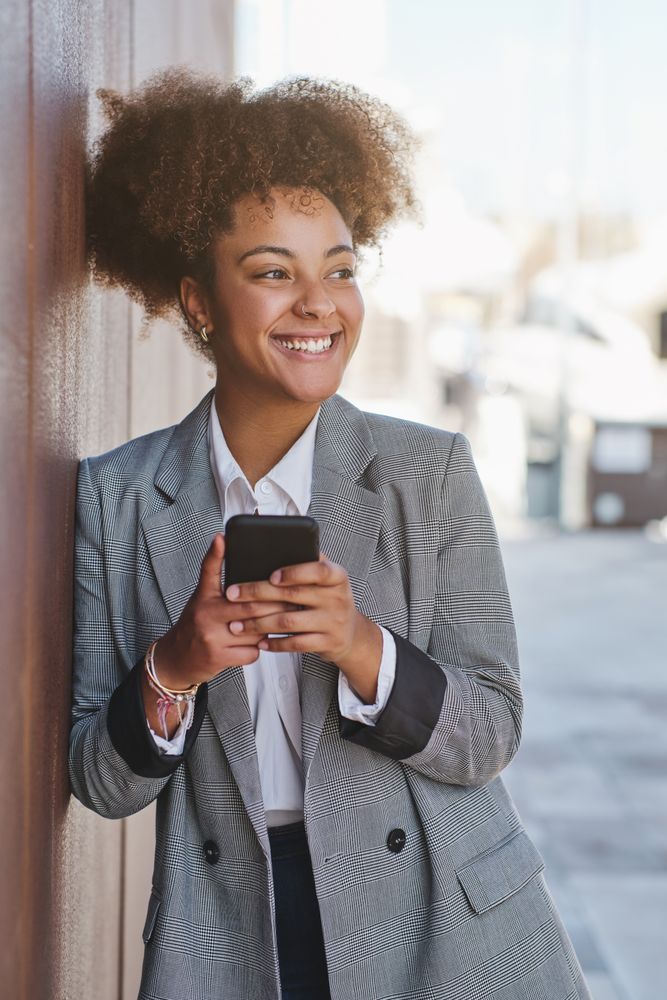 Happy Black woman using phone with smile near street wall
