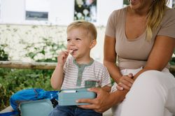 Crop anonymous mother sitting with preschool son with lunch box in daylight