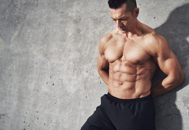 Shirtless muscular serious man leaning against grey wall