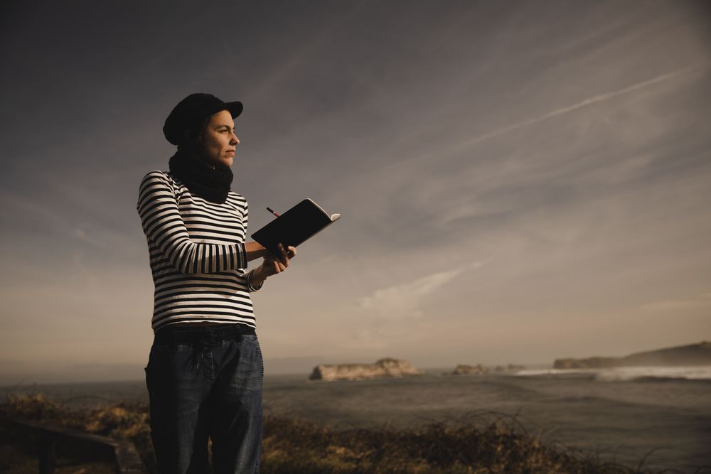 Woman writing in notebook on bench on shore near water