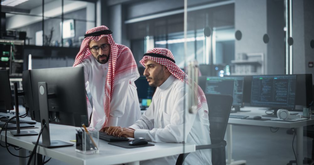 Two Young Saudi Software Engineers Use Desktop Computer to Discuss a Technological Project in a Modern Industrial Office.