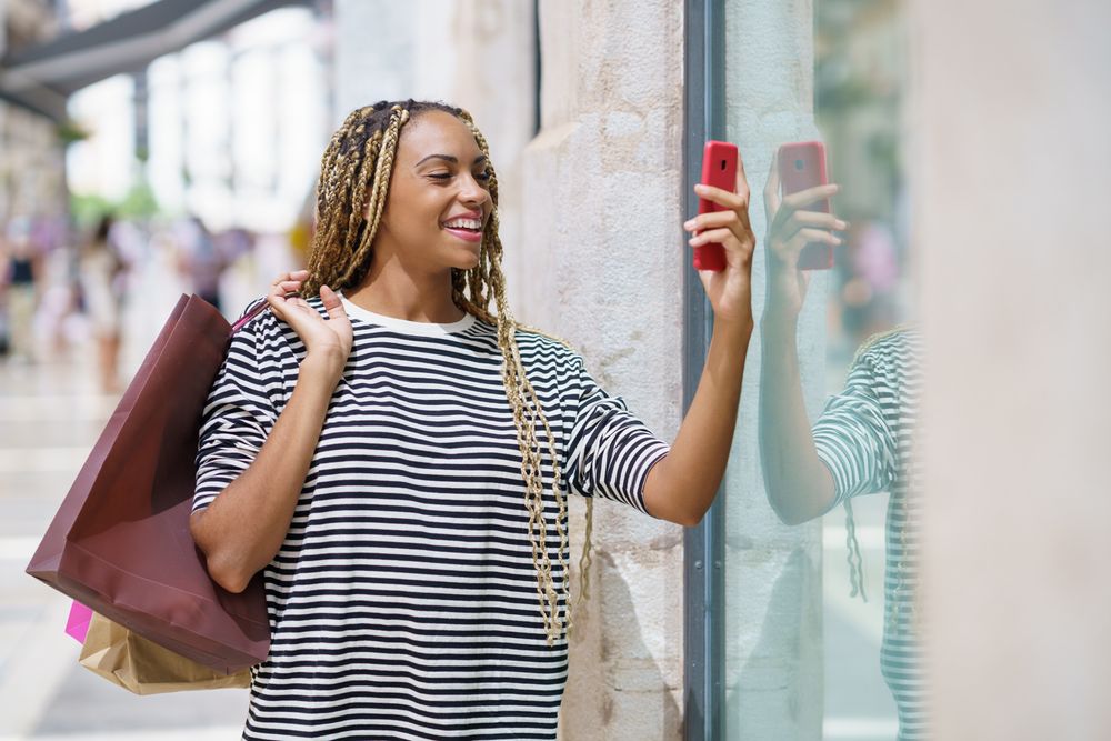 Female with smartphone holding bags over her shoulder reflected through shop window