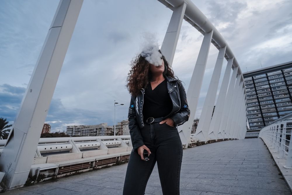 Stylish woman in jacket and jeans smoking on bridge