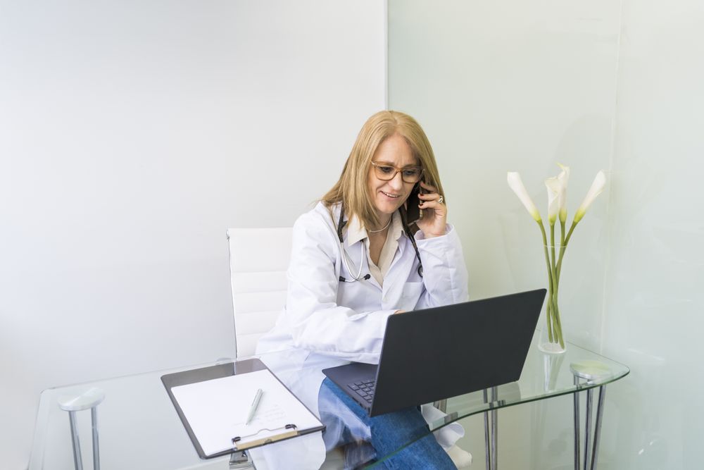 Smiling female doctor talking on the phone to a patient while looking at her medical records on laptop in medical office.
