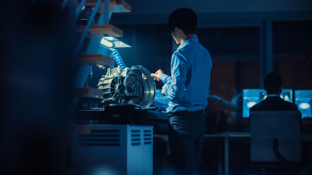 Professional Automotive Engineer in Glasses is Working on Transmission Gears in a High Tech Innovative Laboratory with a Computer Screens.