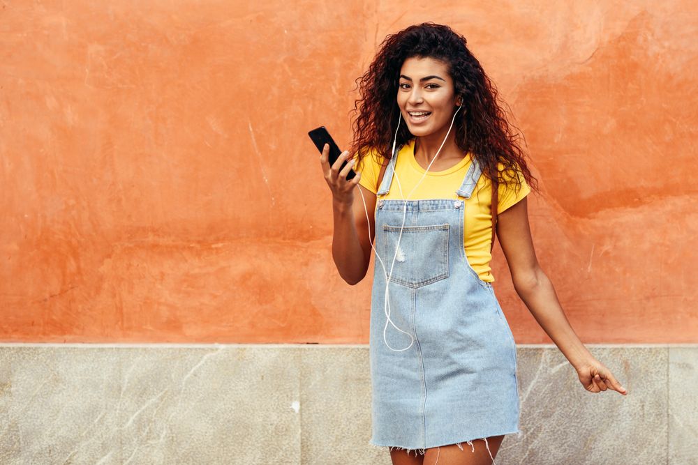 Happy Arab woman with curly hairstyle using phone and headphones in front of on orange wall