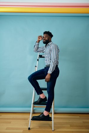 Black man in blue studio standing on step ladder